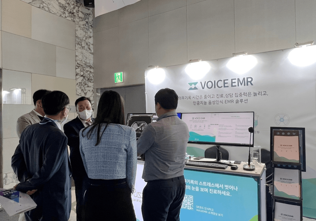 Attendees discussing the VoiceEMR solution at the PuzzleAI booth during the KoSAIM 2022 conference. The booth highlights advanced features related to AI-driven electronic medical records.