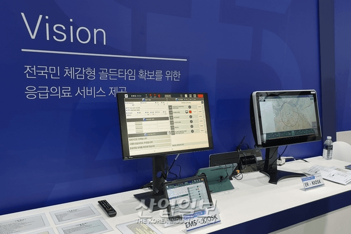 Two computer screens in an emergency medical service kiosk display user interfaces for managing patient information and emergency response operations with a blue backdrop.