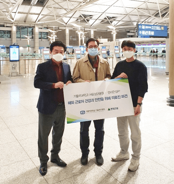 Three individuals at the airport are holding a sign that states, “Medical Dispatch for the Health and Safety of Overseas Workers.” They are wearing masks, highlighting the precautions taken during the COVID-19 pandemic. The setting appears to be an airport terminal, emphasizing the context of their departure to assist overseas workers