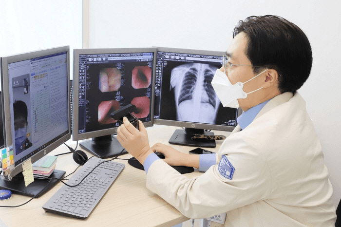 A physician in a white coat wearing a mask is conducting a remote consultation via video call, using dual monitors to review medical imaging results. The left screen displays a video of a patient, while the right showcases various medical images.