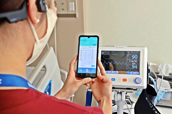 A nurse wearing a mask holds a smartphone displaying the VoiceEMR+ application while monitoring a patient's vital signs on a medical device in a hospital room.