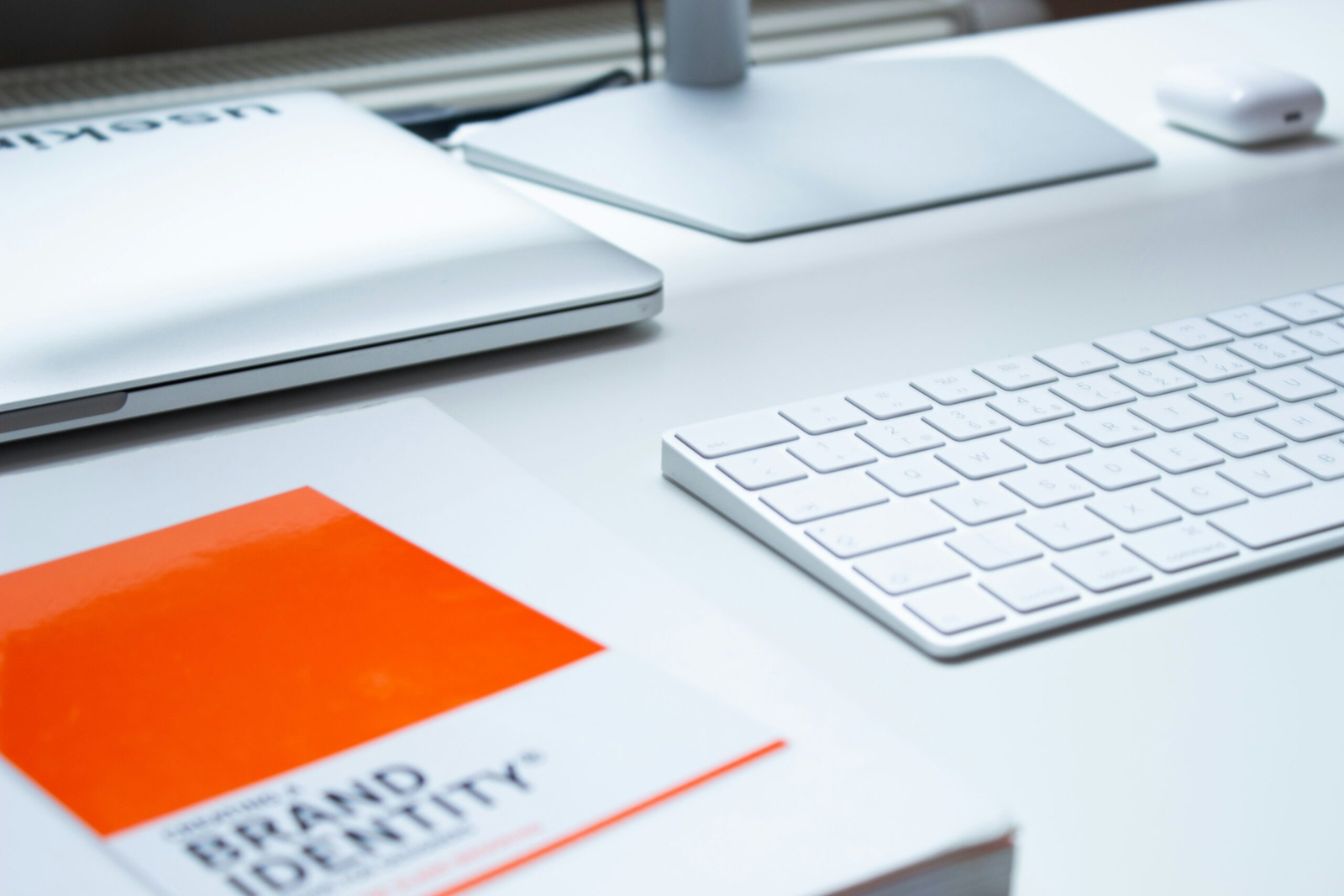 A close-up view of a modern workspace showcasing a sleek silver laptop partially open on a white desk. Beside it is a minimalist white keyboard, and a booklet titled "Brand Identity" with a bold orange cover is prominently displayed. The clean and organized setup highlights a contemporary aesthetic, emphasizing a focus on design and digital tools in a professional environment.