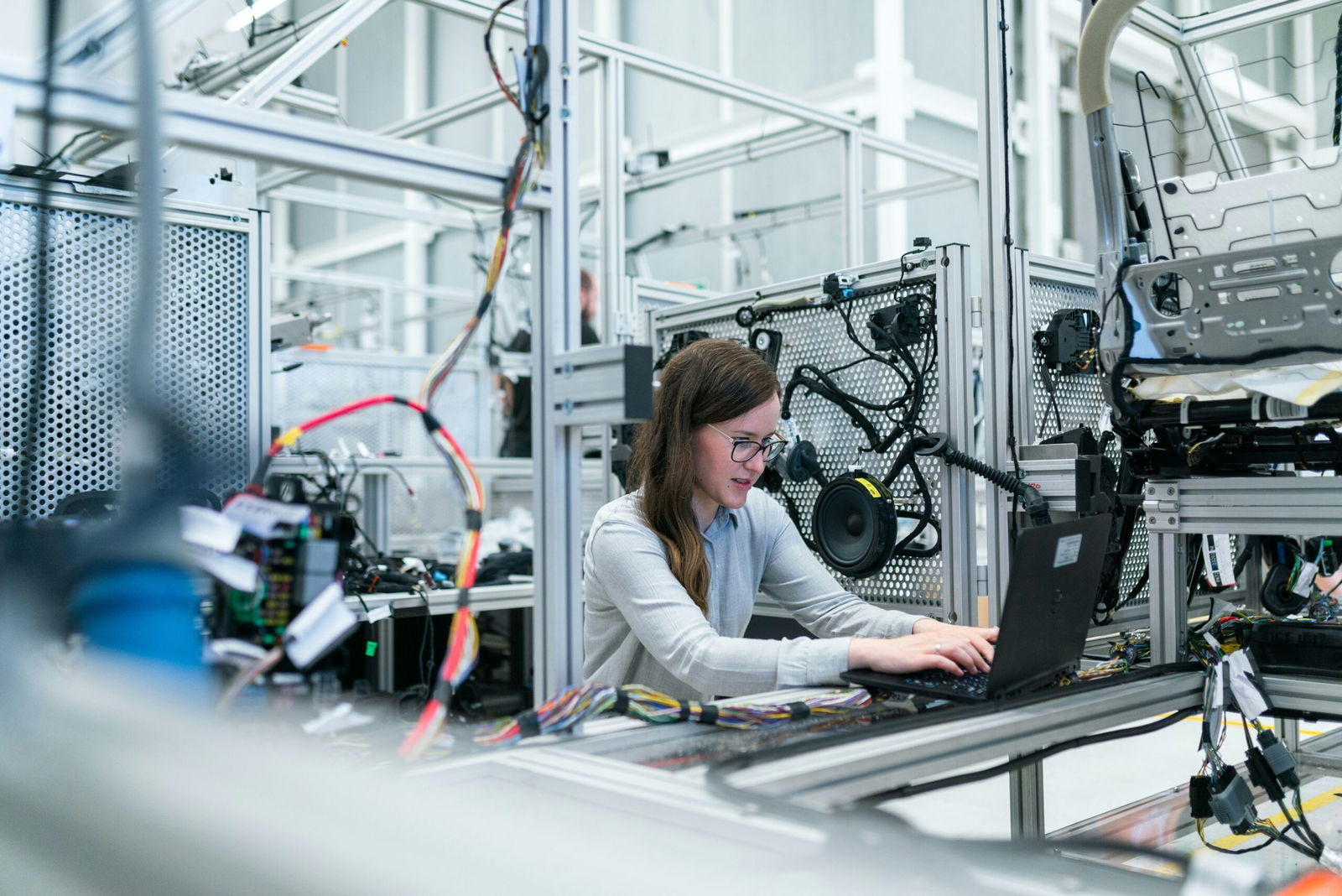 A female engineer with long brown hair and glasses sits at a workstation, focused on her laptop. Surrounding her are various mechanical components and colorful wires, indicating a high-tech environment. The workspace is organized, showcasing the intricate setup of machinery and technical equipment used for engineering tasks.