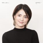 A smiling young woman with short, styled hair poses against a plain background. She is wearing a black turtleneck, and her expression conveys confidence and warmth. A small tattoo is visible on her neck, adding to her unique appearance. The focus is on her friendly demeanor and professional look.